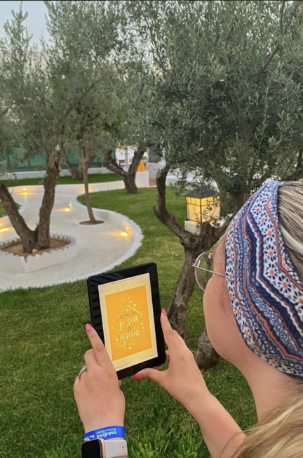 Promis je suis mon héroïne de Camille Roy sur une liseuse, tenue à la main par une jeune femme à lunettes, un bandeau dans les cheveux, dans un espace de verdure avec des oliviers dans un resort en Tunisie