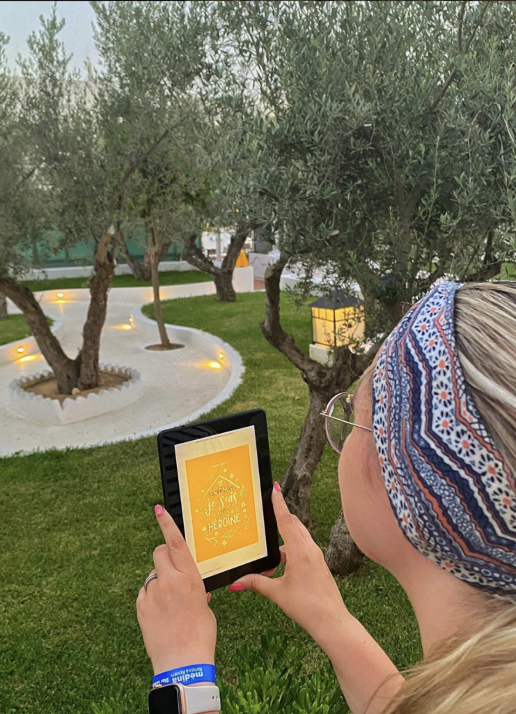 Promis je suis mon héroïne de Camille Roy sur une liseuse, tenue à la main par une jeune femme à lunettes, un bandeau dans les cheveux, dans un espace de verdure avec des oliviers dans un resort en Tunisie