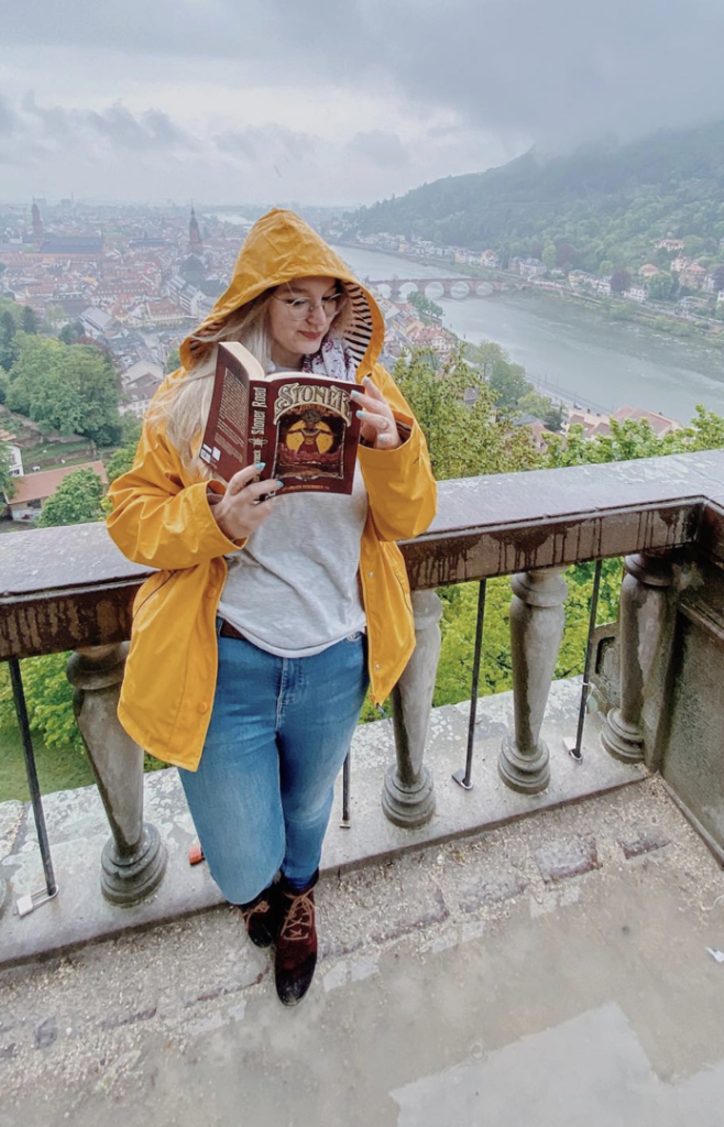 Sorbet-Kiwi en imperméable jaune lisant Stoner Road de Julien Heylbroeck sur un balcon surplombant la ville de Heidelberg enveloppée de brume, évoquant l’esprit road trip et rock du roman.