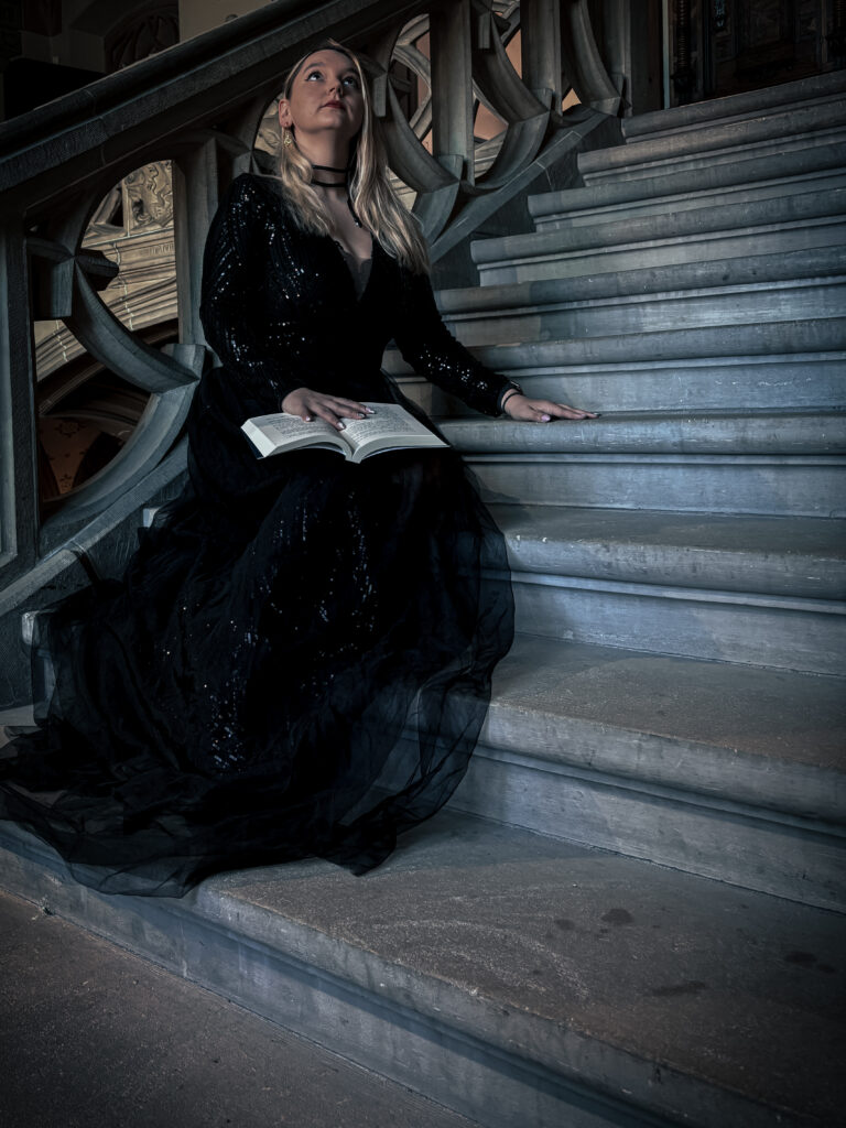 sorbet-Kiwi vêtue d'une longue robe noire pailletée, assise sur l'escalier du Musée historique de Haguenau, un livre ouvert sur les genoux. La lumière tamisée et le décor créent une ambiance mystérieuse.