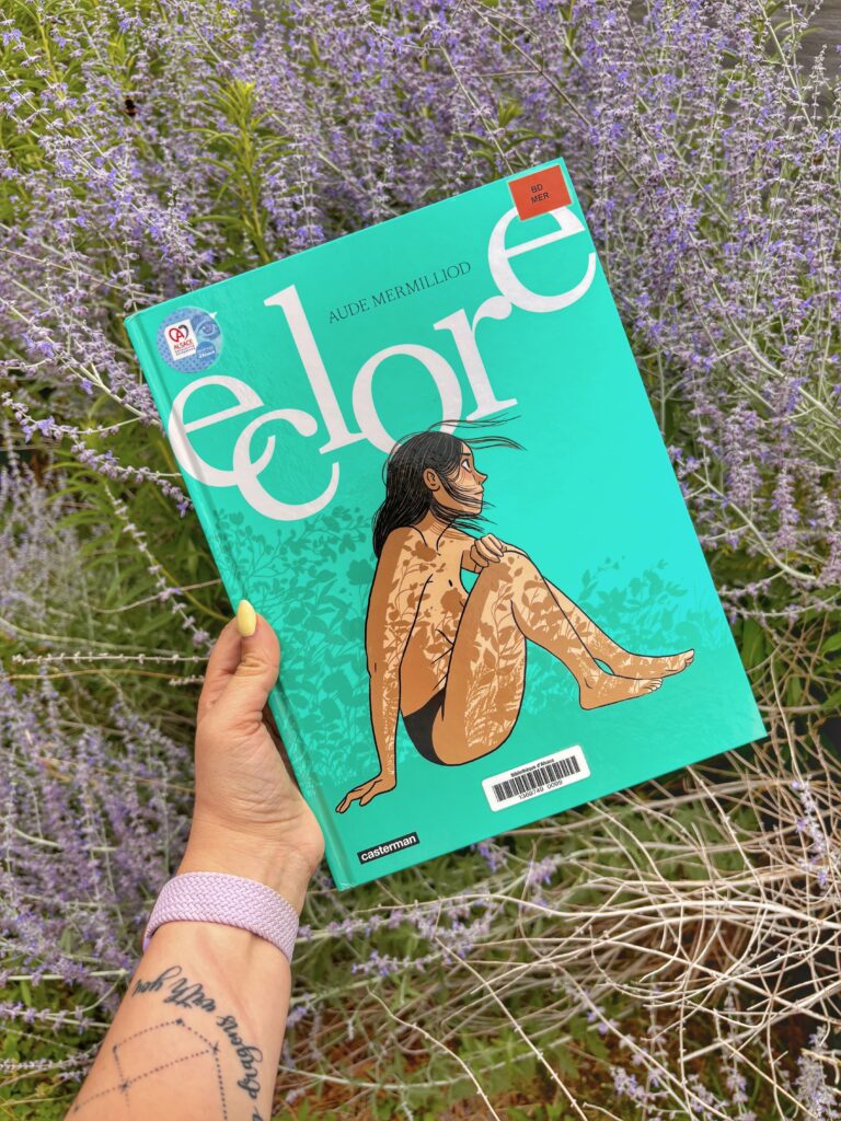 Main tenant la BD éclore d'Aude Mermillod devant un par terre de lavande. La couverture montre une jeune femme assise les genoux relevés sur son torse, juste en petite culotte, des ombres de fleurs se reflétant sur son corps.
