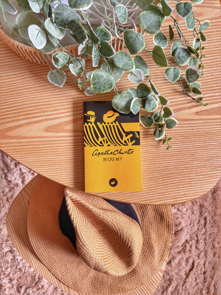 Le roman N ou M d'Agatha Christie posé sur une table gigogne haute avec une plante verte, et sur la gignosse basse un chapeau de paille à l'image de ceux sur la couverture du roman