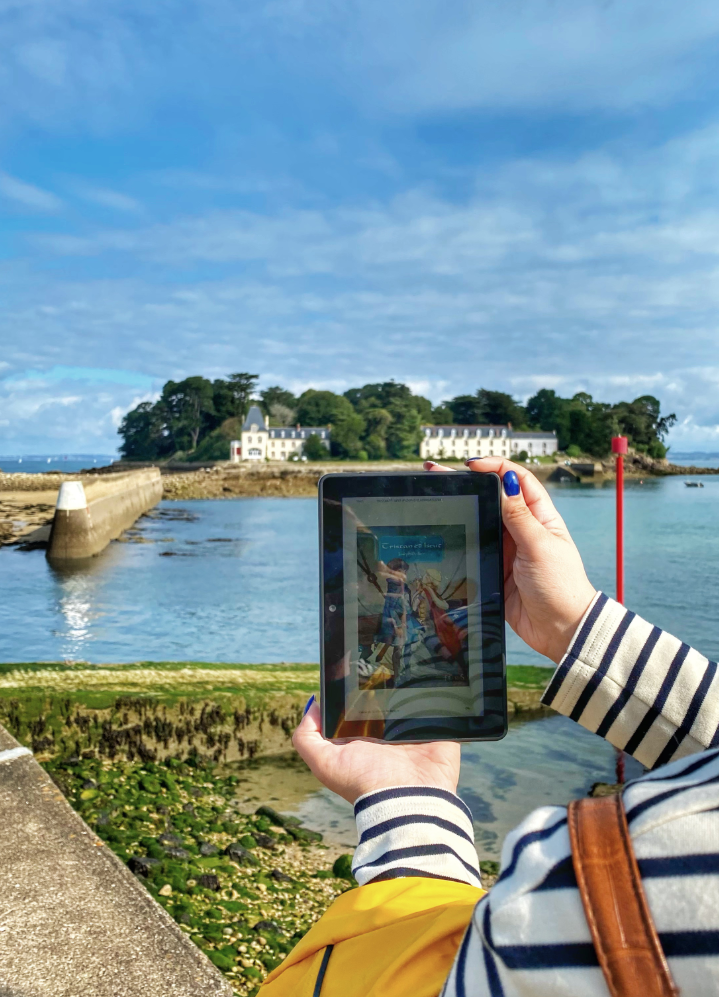Main tenant une kindle avec la couverture de Tristan et Yseut de Joseph Bédier devant l'île Tristan de Douarnenez