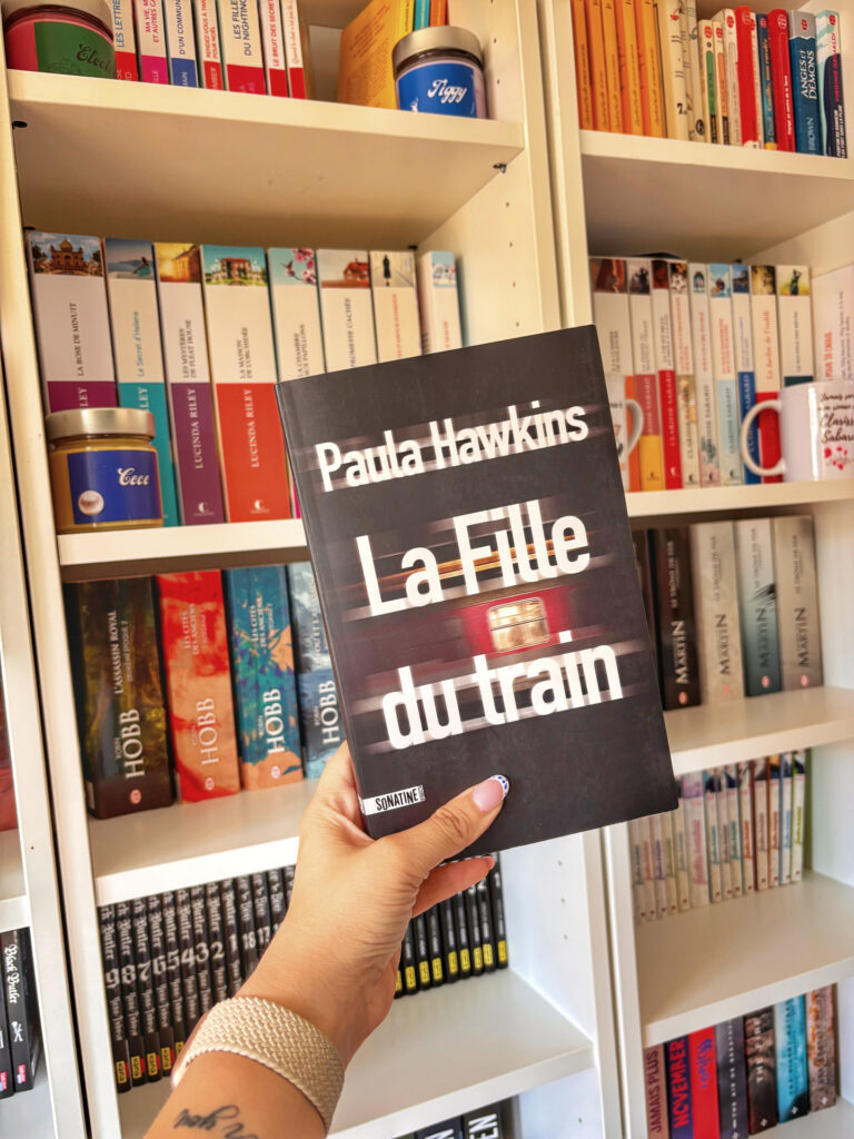Main tenant le roman La fille du train de Paula Hawkins devant une bibliothèque pleine de romans