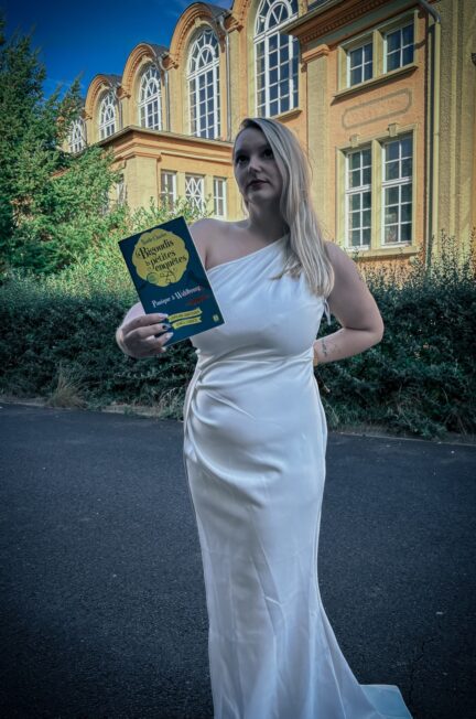 Lecture du roman Panique à Wahlbourg, premier tome de la saga Bigoudis et petites enquêtes de Naëlle Charles, en robe blanche devant un bâtiment historique en Allemagne