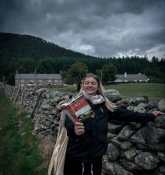 Lecture de La sœur de l’ombre de Lucinda Riley devant un muret de pierres, maisons écossaises en fond sous un ciel menaçant – Loch Lee.