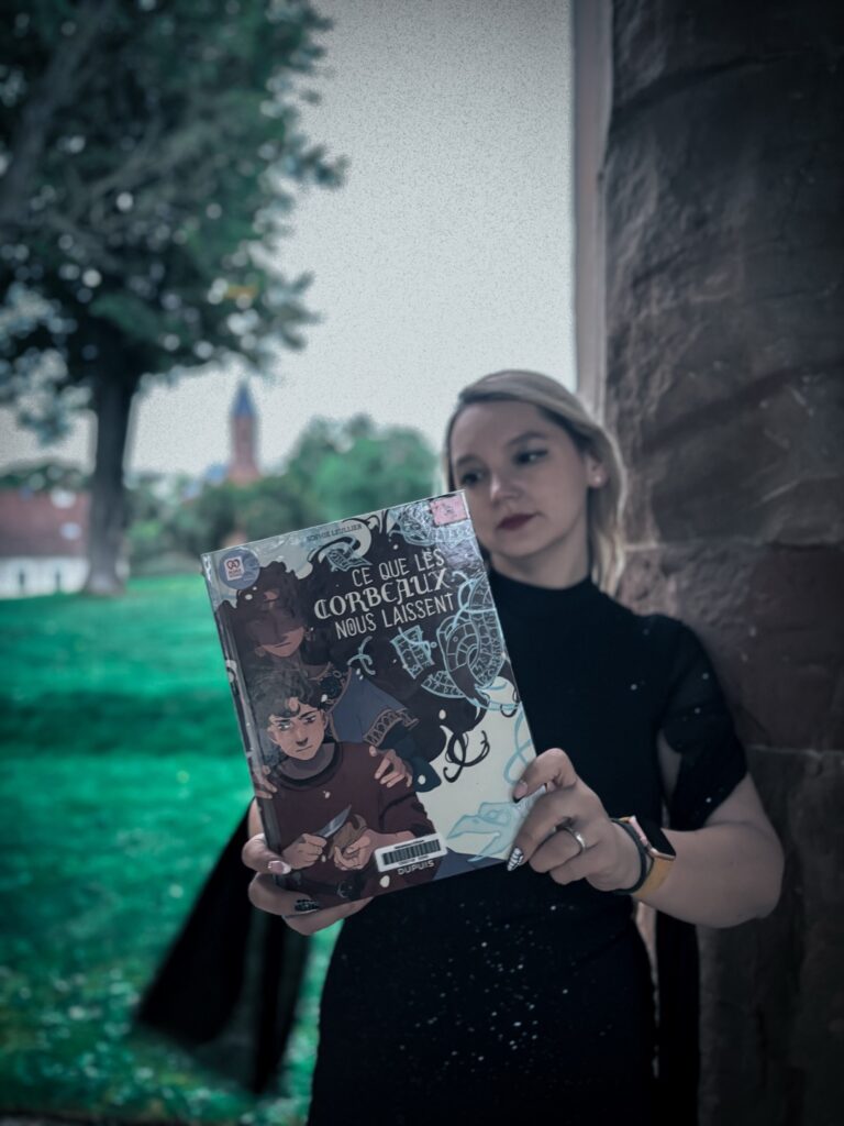 Portrait en robe noire tenant la BD Ce que les corbeaux nous laissent de Sophie Leullier, devant un pilier en pierre et un arbre sombre.