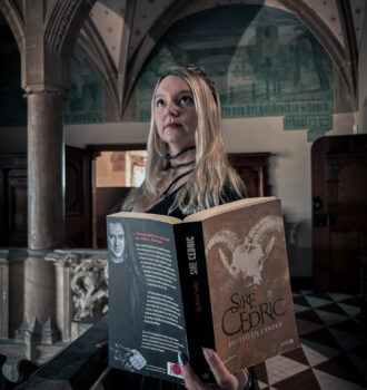 Sorbet-Kiwi en robe noire pailletée lisant le roman Du feu de l’enfer de Sire Cédric dans un bâtiment ancien aux arches gothiques, créant une atmosphère sombre et mystérieuse parfaitement accordée au ton du thriller.