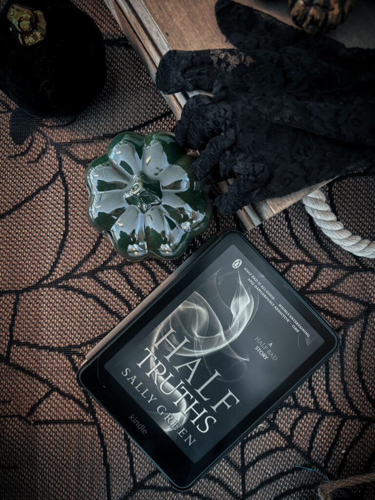 Mise en scène d’Halloween avec une liseuse ouverte sur la couverture de Half Truths de Sally Green, posée sur un tissu à motifs de toiles d’araignée. À côté, des citrouilles décoratives et une paire de gants en dentelle noire accentuent le côté mystérieux et gothique de la composition.