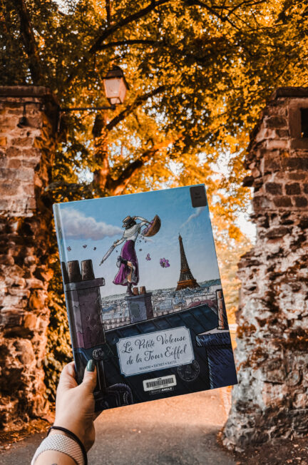 Main tenant la BD La petite voleuse de la Tour Eiffel devant une arche en pierre baignée par la lumière dorée de l’automne, rappelant l’ambiance historique et poétique du récit.