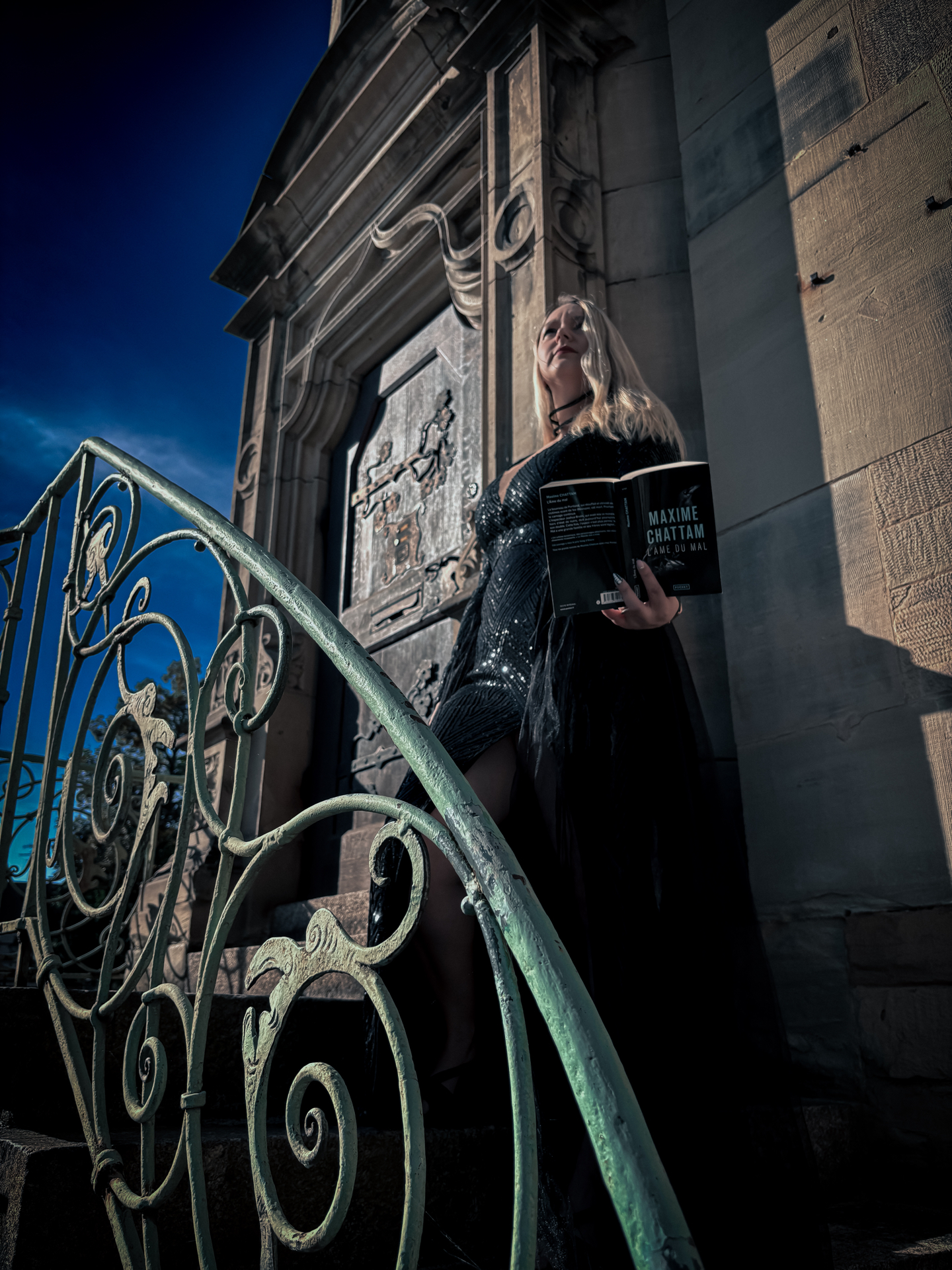 Sorbet-Kiwi pose sur les marchesdu musée historique de Haguenau, vêtue d’une robe noire scintillante, tenant le roman L’âme du mal de Maxime Chattam. L’atmosphère dramatique et les ombres renforcent l’ambiance sombre du thriller.