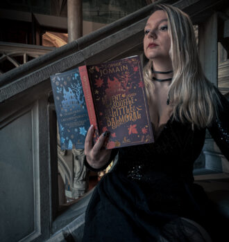 Sorbet-Kiwi en robe noire pailletée lisant le roman Le vent souffle sur Little Balmoral de Sophie Jomain, posée sur un escalier ancien.