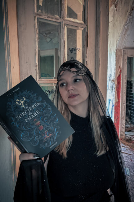 Sorbet-Kiwi pose dans une bâtisse abandonnée, vêtue d’une robe noire et d’un voile sombre, tenant le roman Les sorcières du phare de C. J. Cooke. L’ambiance mystérieuse et légèrement gothique rappelle l’atmosphère du livre et la saison d’Halloween.