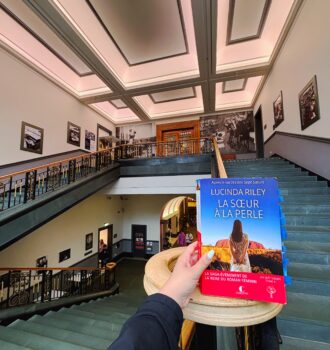 Photo d’une main tenant le roman La sœur à la perle de Lucinda Riley dans l’entrée monumentale de la Bibliothèque nationale d’Écosse, avec son grand escalier et son architecture intérieure lumineuse.