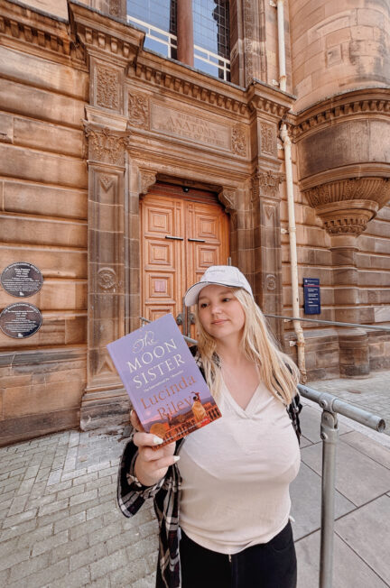 photo de Sorbet-Kiwi tenant le roman The Moon Sister de Lucinda Riley devant la porte principale en pierre de la Faculté de médecine d’Édimbourg, surmontée de l’inscription “Anatomy and Practice of Physic”.