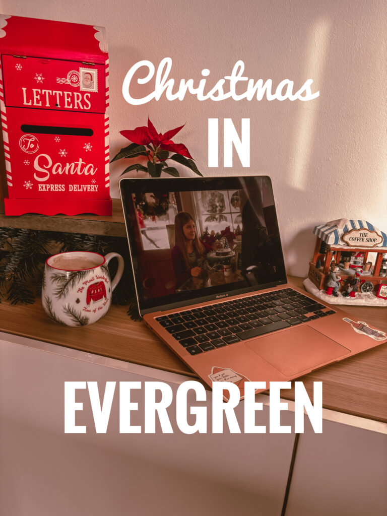 Un ordinateur portable posé sur un meuble diffuse une scène du premier Christmas in Evergreen. À côté, une tasse décorée, une guirlande de sapin et une petite maison de village enneigé sont disposées. Sur la gauche, une grande boîte rouge en forme de boîte aux lettres indique Letters to Santa.