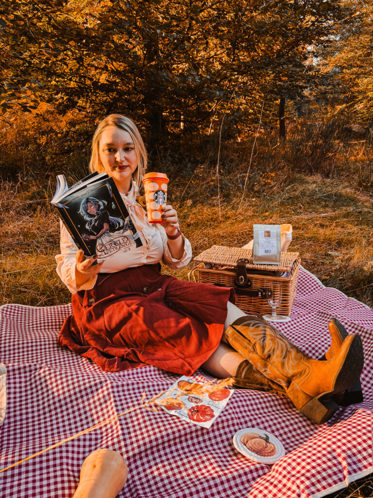 Sorbet-Kiwi assise sur une nappe vichy rouge en clairière d'automne, lisant Le cercle de l'étrange 2 et tenant un gobelet orange Starbucks, panier de pique-nique à coté, avec des bottes western dans une lumière dorée.