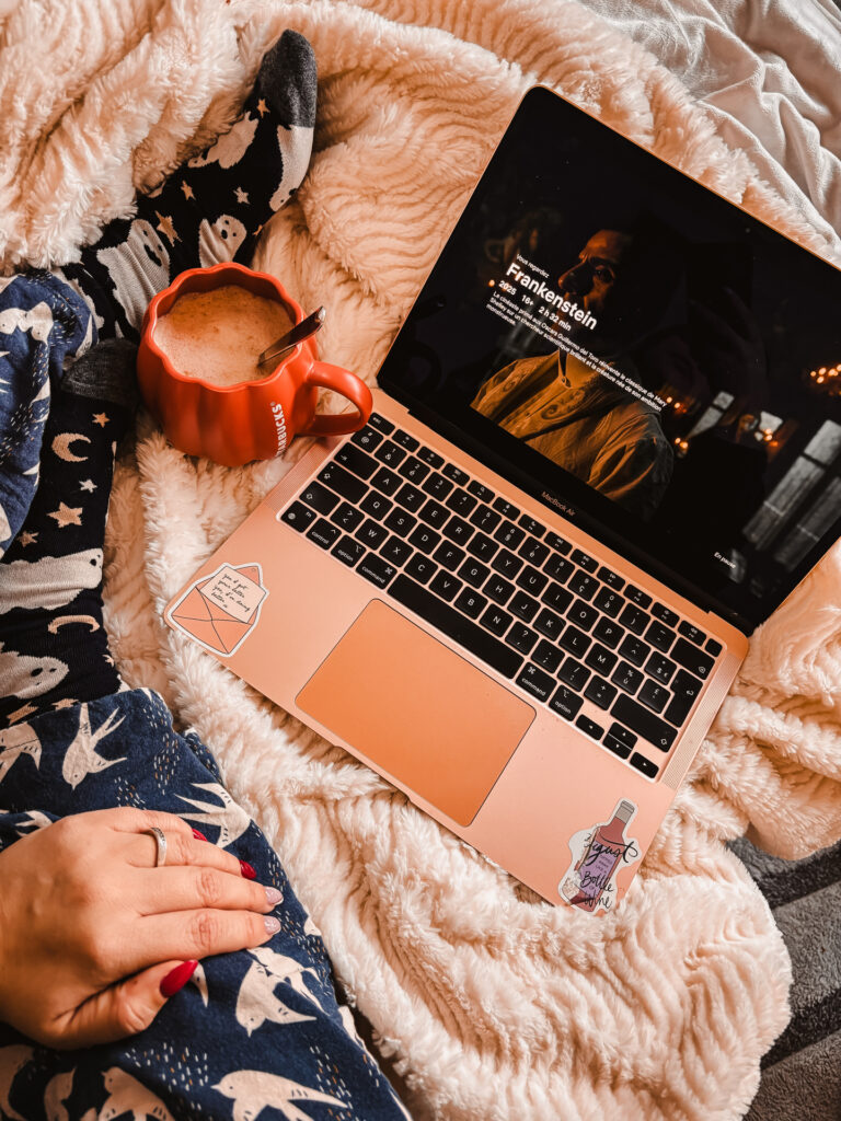 Sorbet-Kiwi en pyjama bleu imprimé d’hirondelles est installée sur un plaid blanc duveteux, devant un ordinateur portable rose affichant la page du film Frankenstein sur Netflix. À gauche, une tasse citrouille orange remplie d’une boisson chaude repose près de ses pieds en chaussettes décorées de fantômes et de lunes. L’ambiance est cosy, automnale et chaleureuse.
