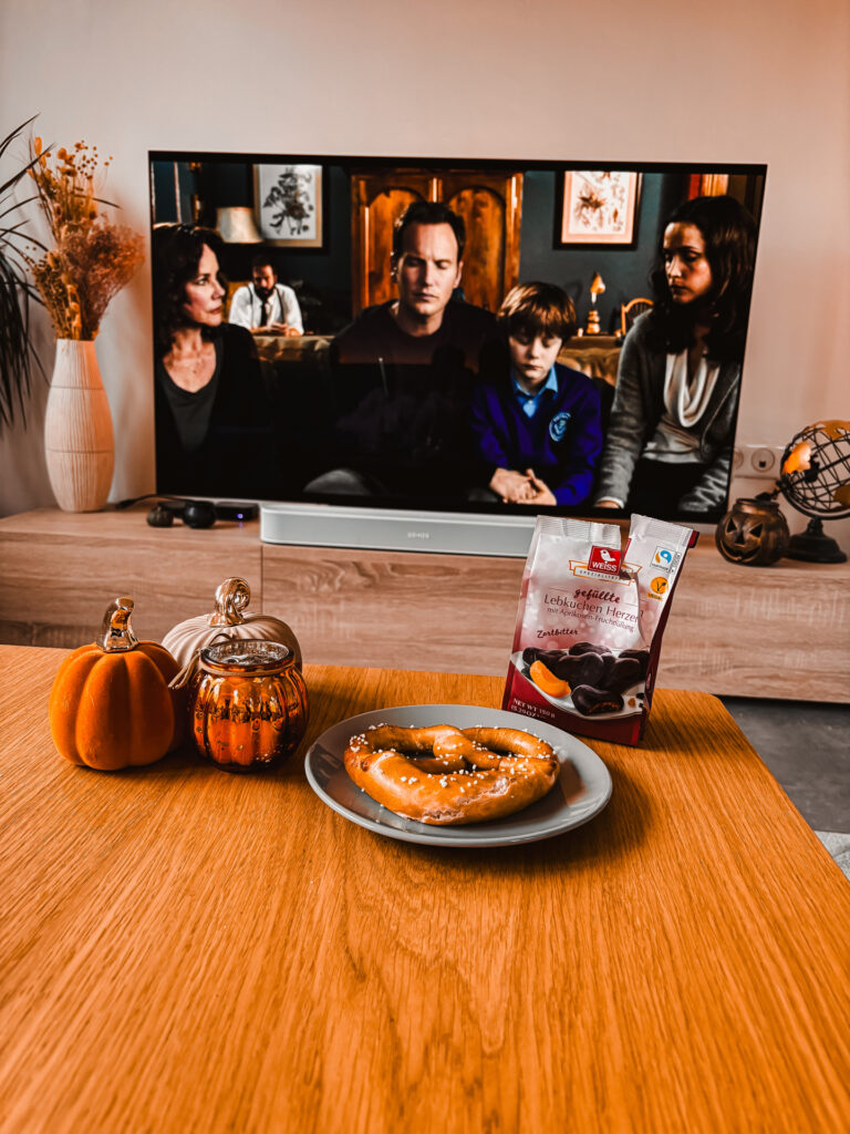 Salon décoré aux couleurs de l’automne avec des citrouilles et des pâtisseries, un écran diffusant une scène du film Insidious : The Red Door, créant une ambiance entre frisson et cocooning typique de la saison Noëlloween.