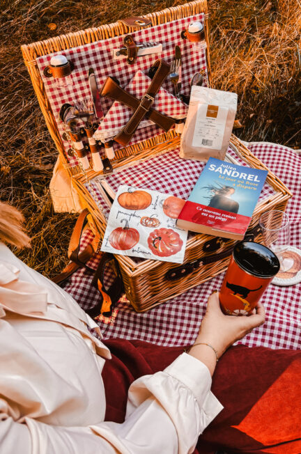 Scène automnale de pique-nique littéraire, avec un panier en osier, un thermos orangé Starbucks et le roman Le jour où Rose a disparu de Julien Sandrel posé sur une nappe à carreaux rouges, dans une lumière dorée de fin de journée.