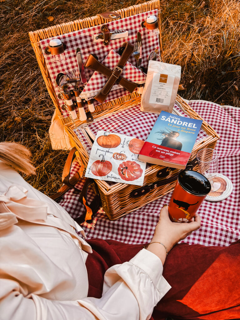 Scène automnale de pique-nique littéraire, avec un panier en osier, un thermos orangé Starbucks et le roman Le jour où Rose a disparu de Julien Sandrel posé sur une nappe à carreaux rouges, dans une lumière dorée de fin de journée.