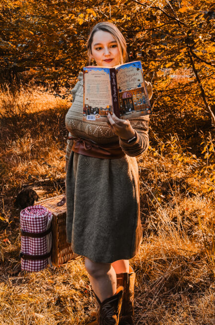 Sorbet-Kiwi en robe et bottes automnales lisant le roman Le Pumpkin Spice Café de Laurie Gilmore en pleine nature dorée, avec un panier de pique-nique et une couverture à carreaux, dans une lumière chaude évoquant l’esprit cosy et sucré de l’automne.