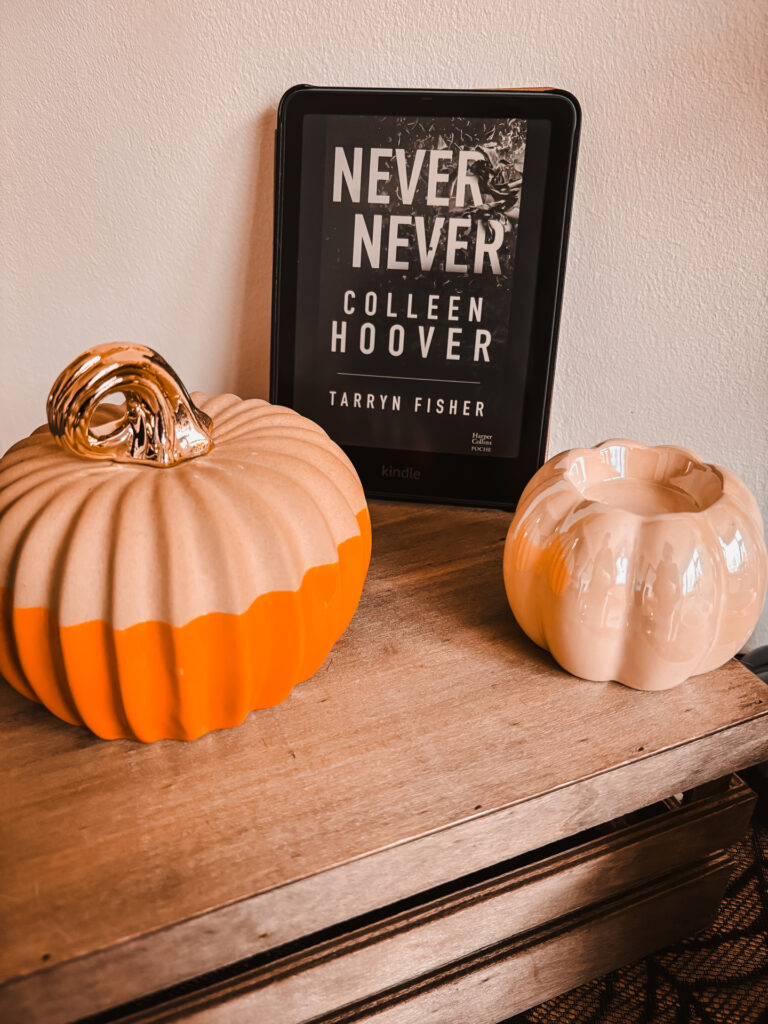 Table en bois décorée de deux citrouilles céramiques et d’une liseuse Kindle affichant la couverture du roman Never Never de Colleen Hoover et Tarryn Fisher, dans une ambiance automnale douce et chaleureuse.