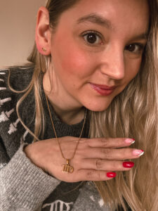 Un gros plan montre Sorbet-Kiwi portant des boucles d’oreilles dorées et un collier assorti avec un pendentif en forme de lettres TTPD. Sa main droite montre une manucure rouge et rose. - Cadeaux pour Swifties