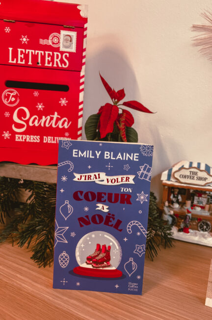 Un exemplaire du roman J’irai voler ton cœur à Noël est posé sur un meuble en bois. À gauche, une boîte aux lettres décorative rouge indique Letters to Santa. À droite, une petite maison de village de Noël représente un coffee shop. Une plante poinsettia se trouve derrière le livre.