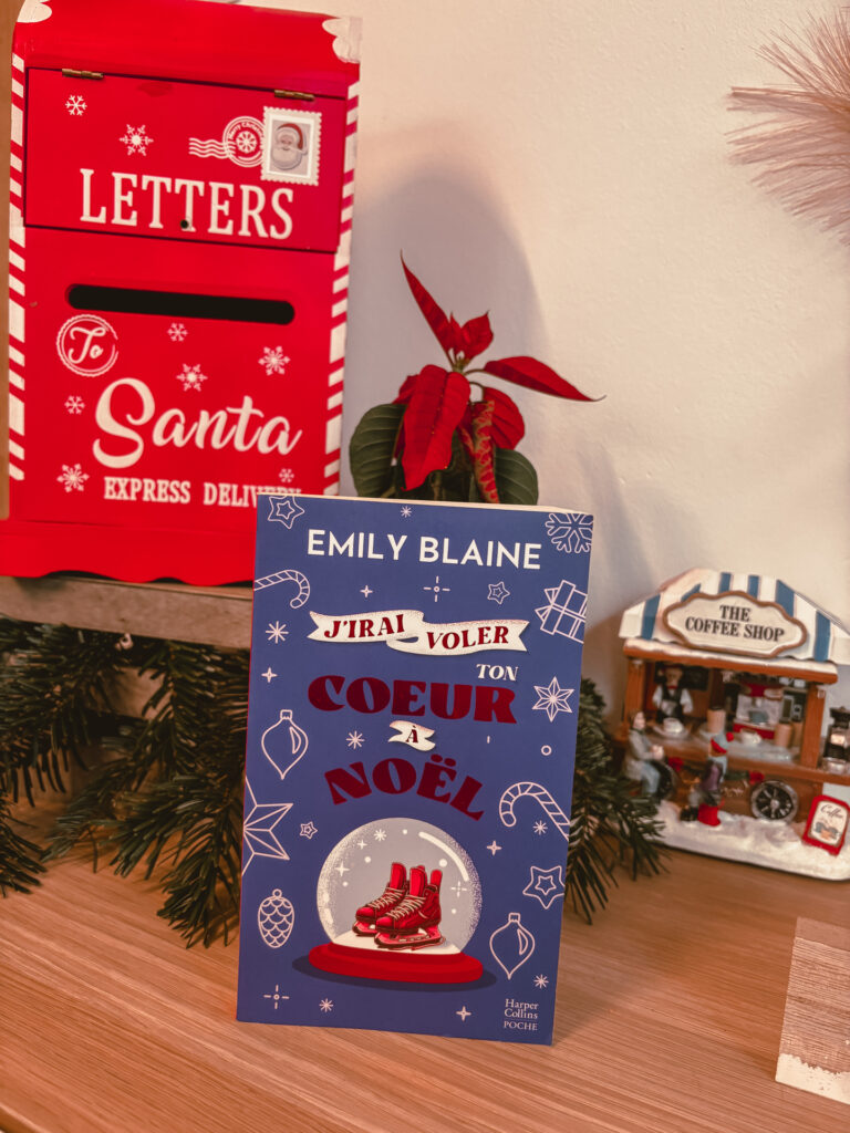 Un exemplaire du roman J’irai voler ton cœur à Noël est posé sur un meuble en bois. À gauche, une boîte aux lettres décorative rouge indique Letters to Santa. À droite, une petite maison de village de Noël représente un coffee shop. Une plante poinsettia se trouve derrière le livre.