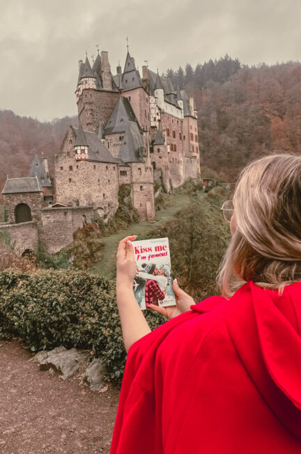 Sorbet-Kiwivêtue d’un manteau rouge tient le livre Kiss Me I’m Famous devant le château médiéval d’Château d’Eltz, en Allemagne, lors d’une séance photo de mariage par temps d’automne.