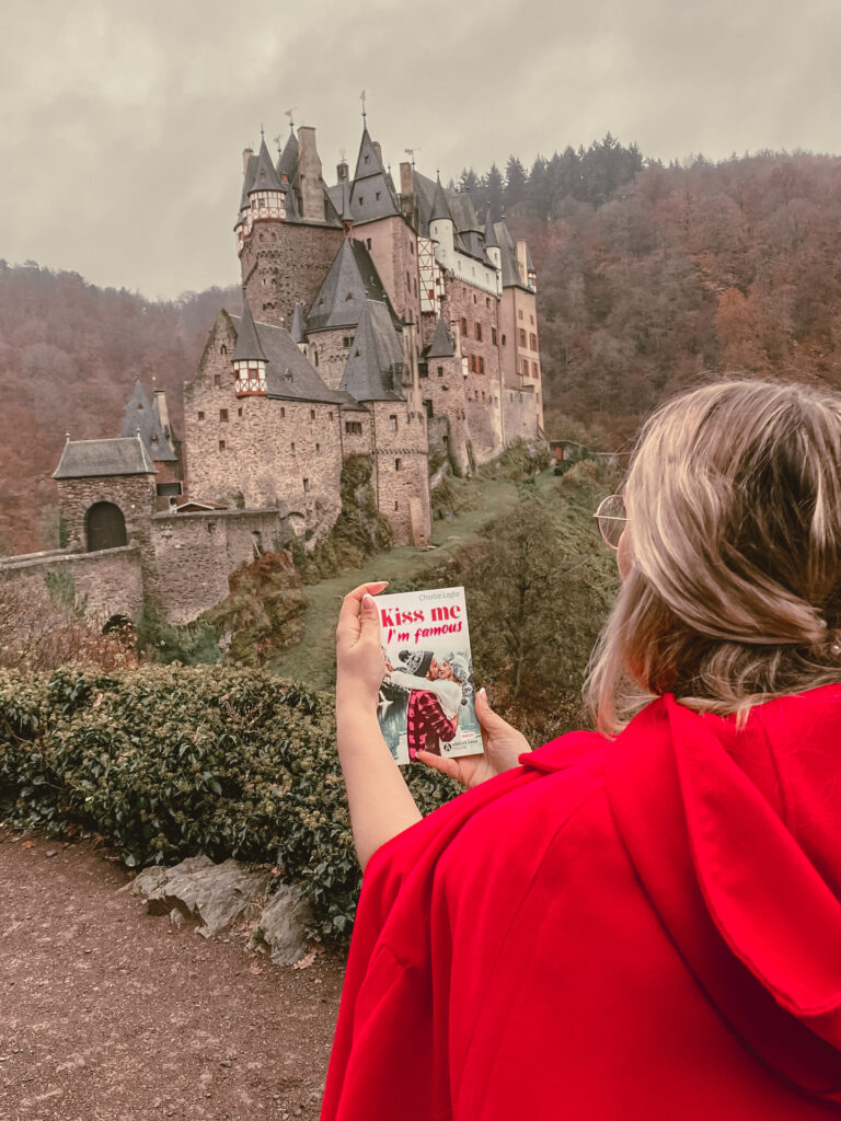 Sorbet-Kiwivêtue d’un manteau rouge tient le livre Kiss Me I’m Famous devant le château médiéval d’Château d’Eltz, en Allemagne, lors d’une séance photo de mariage par temps d’automne.