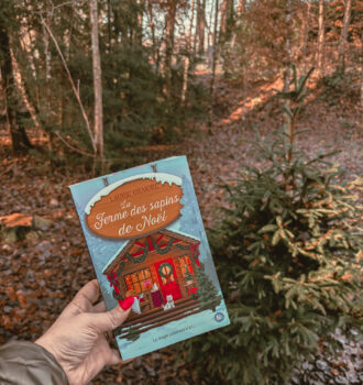 Le roman La ferme des sapins de Noël de Laurie Gilmore tenu à la main en pleine forêt vosgienne, avec des sapins et un sous-bois aux couleurs d’automne en arrière-plan.
