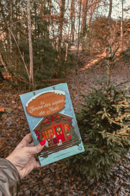 Le roman La ferme des sapins de Noël de Laurie Gilmore tenu à la main en pleine forêt vosgienne, avec des sapins et un sous-bois aux couleurs d’automne en arrière-plan.