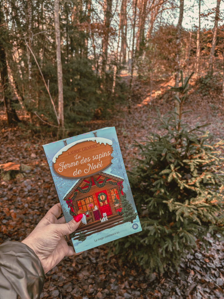Le roman La ferme des sapins de Noël de Laurie Gilmore tenu à la main en pleine forêt vosgienne, avec des sapins et un sous-bois aux couleurs d’automne en arrière-plan.