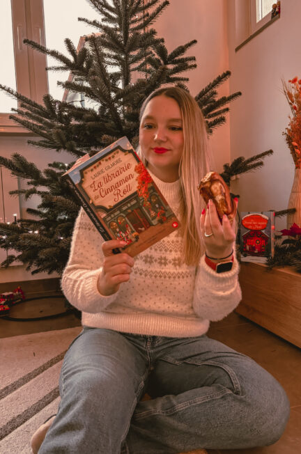 Sorbet-Kiwi assise au sol devant un sapin de Noël tient le roman La librairie du Cinnamon Roll. Elle porte un pull blanc à motifs de Noël et un jean. Dans l’autre main, elle tient une cinnamon roll. Des décorations de Noël et un petit train sont visibles autour d’elle.