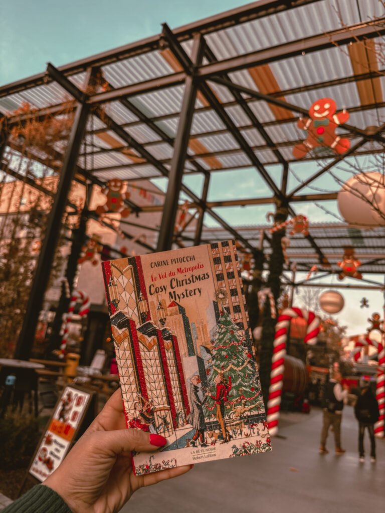 Une main tient le roman Le vol du Metropolis devant une allée décorée pour Noël. Des cannes d’orchidée rouges et blanches, des guirlandes et des silhouettes en pain d’épices sont suspendues. Une structure métallique avec toit transparent apparaît en arrière-plan. La photo a été prise à Shopping Promenade Mundolsheim.