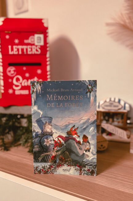 Le roman L’esprit de l’hiver – Mémoires de la forêt, tome 3 de Mickaël Brun-Arnaud, illustré par Sanoe, est posé sur une étagère en bois. En arrière-plan, une boîte aux lettres rouge décorative de Noël et un petit décor hivernal créent une ambiance chaleureuse et festive.