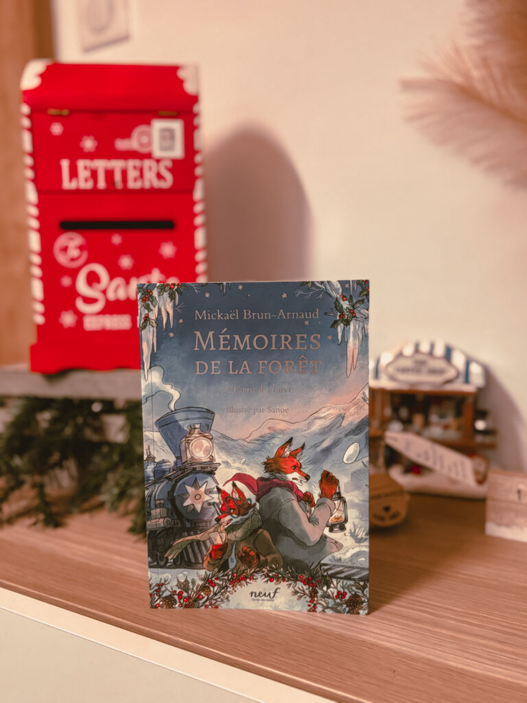 Le roman L’esprit de l’hiver – Mémoires de la forêt, tome 3 de Mickaël Brun-Arnaud, illustré par Sanoe, est posé sur une étagère en bois. En arrière-plan, une boîte aux lettres rouge décorative de Noël et un petit décor hivernal créent une ambiance chaleureuse et festive.