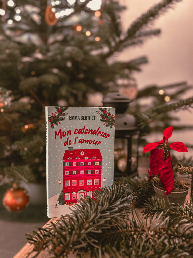 Livre Mon calendrier de l’amour d’Emma Berthet posé devant un sapin décoré, entouré de branches de sapin et d’un poinsettia rouge, dans une ambiance chaleureuse de Noël.