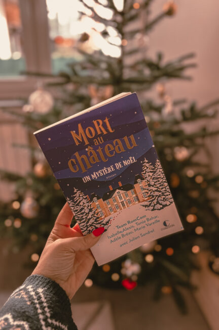Le livre Mort au château : Un mystère de Noël tenu à la main devant un sapin de Noël décoré, avec des lumières floues en arrière-plan.