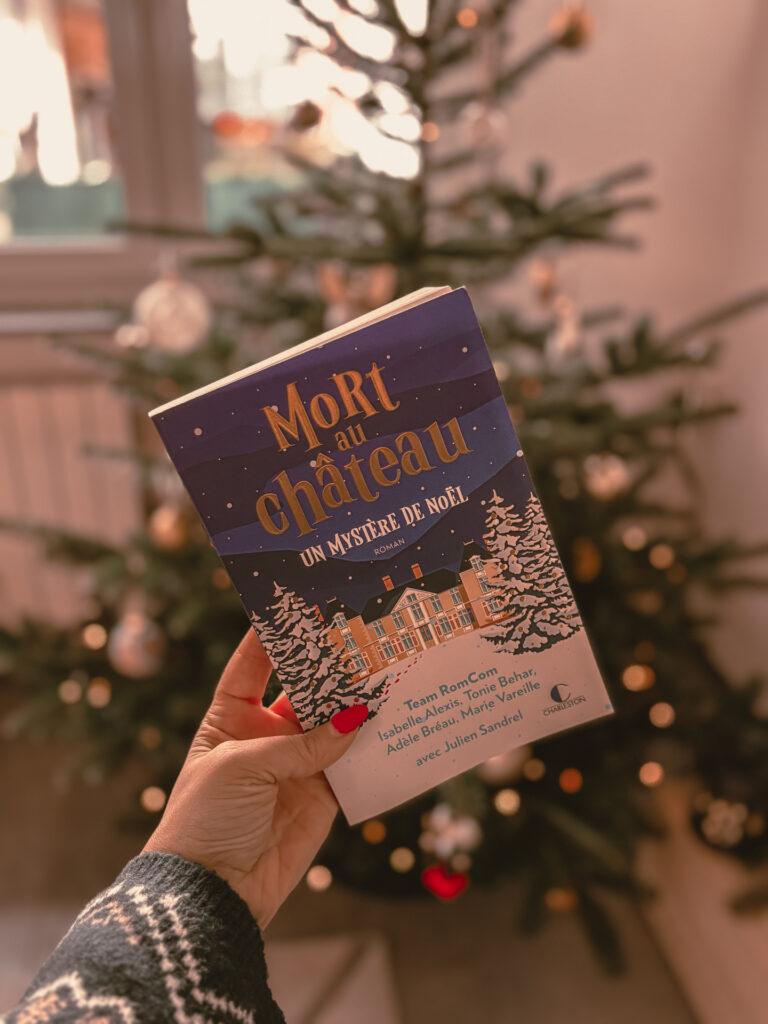Le livre Mort au château : Un mystère de Noël tenu à la main devant un sapin de Noël décoré, avec des lumières floues en arrière-plan.