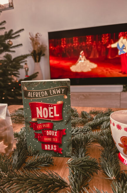 Un exemplaire du roman Noël est plus doux dans tes bras est posé sur une table entourée de branches de sapin. À gauche, un sachet cadeau doré avec des étoiles à la cannelle dedans et à droite une tasse décorée avec un bonhomme pain d’épice. En arrière-plan, un sapin décoré et une télévision diffusant une scène du film Anastasia.