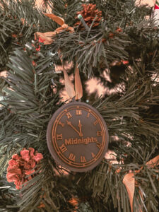 Une décoration en forme d’horloge portant l’inscription “Midnights” est accrochée dans un sapin artificiel. On voit des rubans dorés et des pommes de pin autour. - Cadeaux pour Swifties