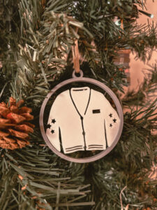 Une décoration en forme de cardigan est accrochée dans un sapin artificiel. On voit des rubans dorés et des pommes de pin autour. - Cadeaux pour Swifties