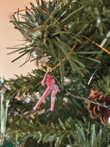 Une petite silhouette découpée représentant Taylor Swift en bodysuit rose est suspendue dans un sapin artificiel. La branche comporte aussi des pommes de pin. - Cadeaux pour Swifties