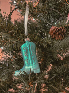 Une botte décorative turquoise suspendue à une branche de sapin artificiel. Des motifs pailletés apparaissent sur l’ornement, entouré d’aiguilles vertes. - Cadeaux pour Swifties