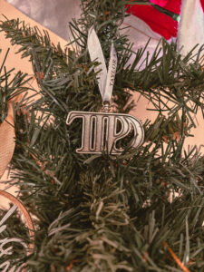 Une décoration en forme de lettres portant l’inscription “TTPD” est accrochée dans un sapin artificiel. On voit des rubans dorés et des pommes de pin autour. - Cadeaux pour Swifties