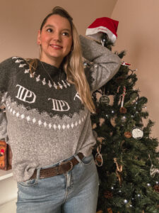 Sorbet-Kiwi porte un pull gris de la merch officielle Taylor Swift de la collection TTPD décoré de motifs blancs de Noël, pose une main derrière la tête et se tient devant un sapin de Noël. Un bonnet rouge repose sur la cime du sapin. Cadeaux pour Swifties