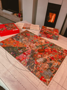 Un grand puzzle coloré représentant des éléments associés à Taylor Swift est posé sur une table. La boîte et un livret explicatif sont ouverts à côté. Une cheminée allumée se trouve en arrière-plan.